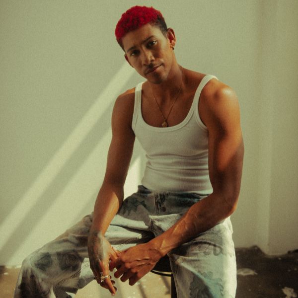 Keiynan Lonsdale sitting on stool in denim jeans and white singlet with short red crew cut hair