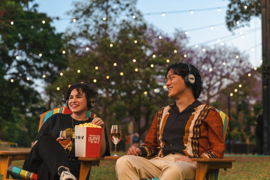 Two people sit in outdoor chairs under string lights at the Dendy Powerhouse Outdoor Cinema, smiling and holding drinks. A popcorn bucket rests between them. Trees and a twilight sky frame the scene at Brisbane Powerhouse, creating a cozy atmosphere.