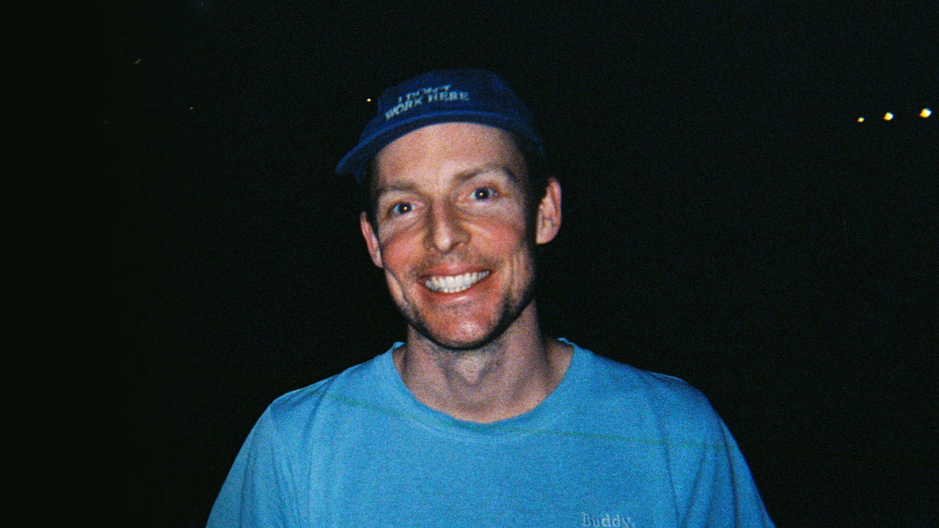 A person wearing a blue shirt and a blue cap with text smiles in a dimly lit setting. The dark background highlights the performer's face and attire, capturing the festive vibe.