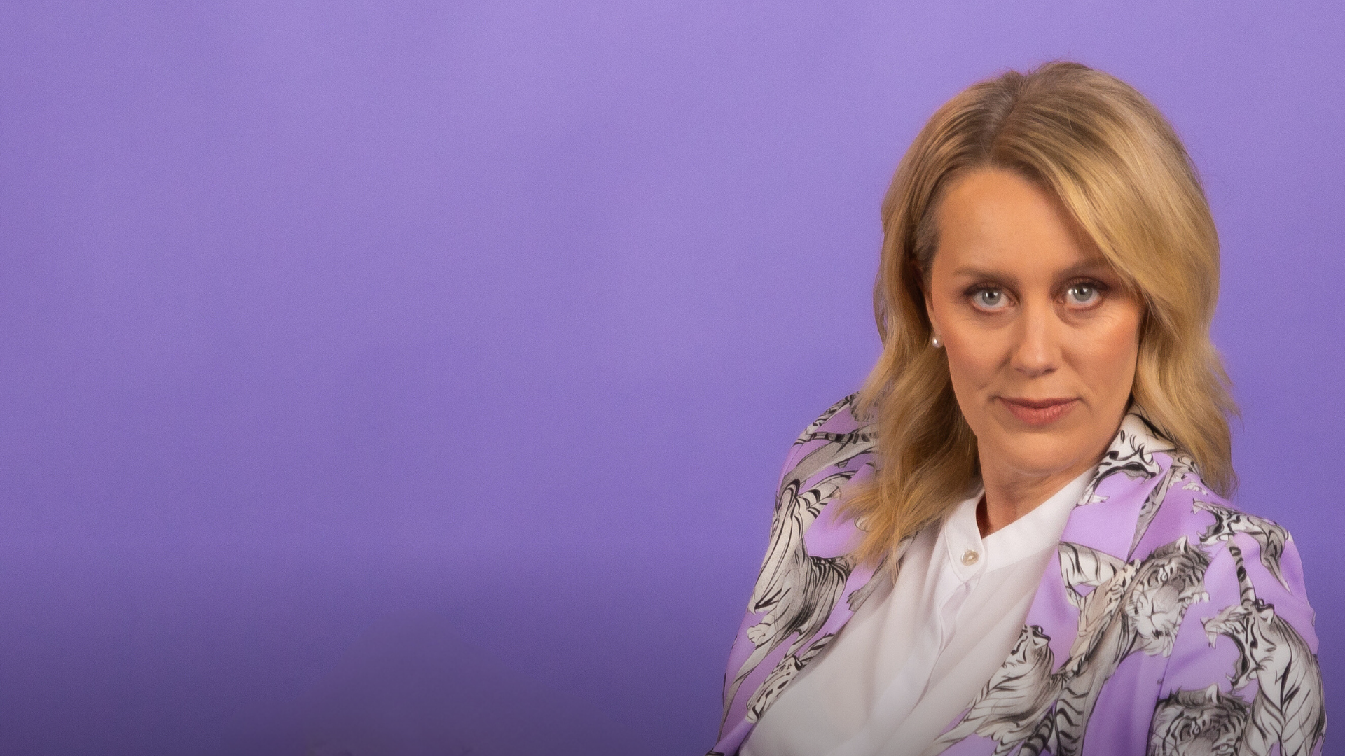 Claire Hooper, set to dazzle at the Brisbane Comedy Festival 2025, sits against a solid purple background. With her blonde hair and floral-patterned purple blazer over a white top, she looks directly at the camera with a neutral expression, capturing her comedic poise before taking the Powerhouse stage.