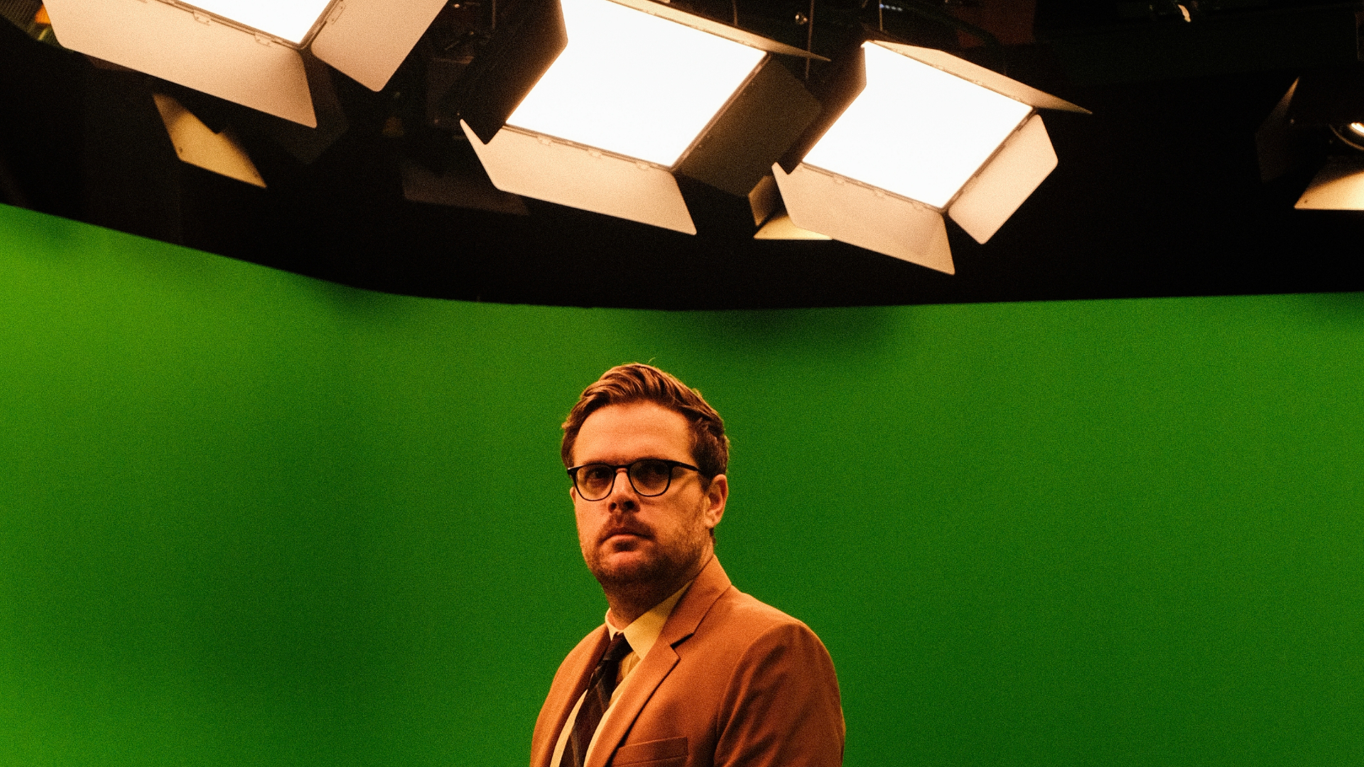 A man in a brown suit stands in front of a green screen, illuminated by studio lights above. He's wearing glasses and has a serious expression. The setting suggests a film or television production studio.