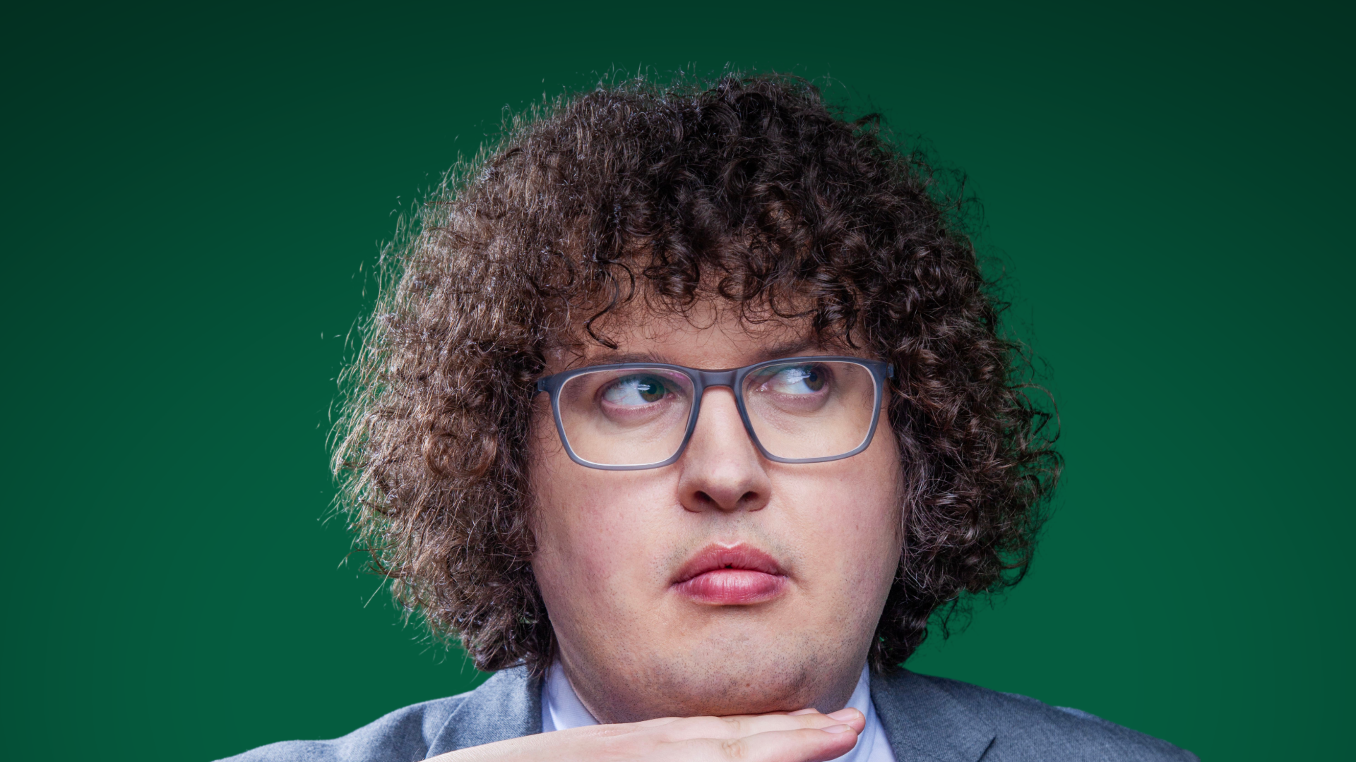 A person with curly hair and glasses, gazes pensively to the side against a green backdrop. They wear a suit and rest their chin thoughtfully on their hand.