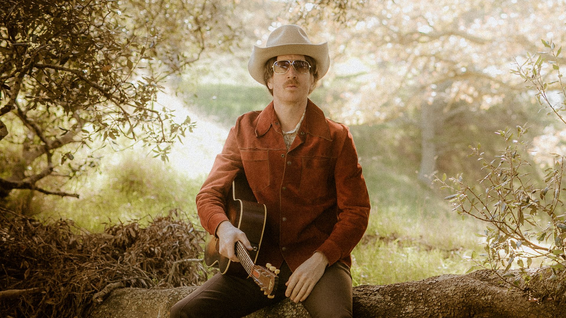 A person wearing a cowboy hat, sunglasses, and a red jacket sits on a rock holding an acoustic guitar, amid the lush greenery, with sunlight filtering through the leaves like spotlights