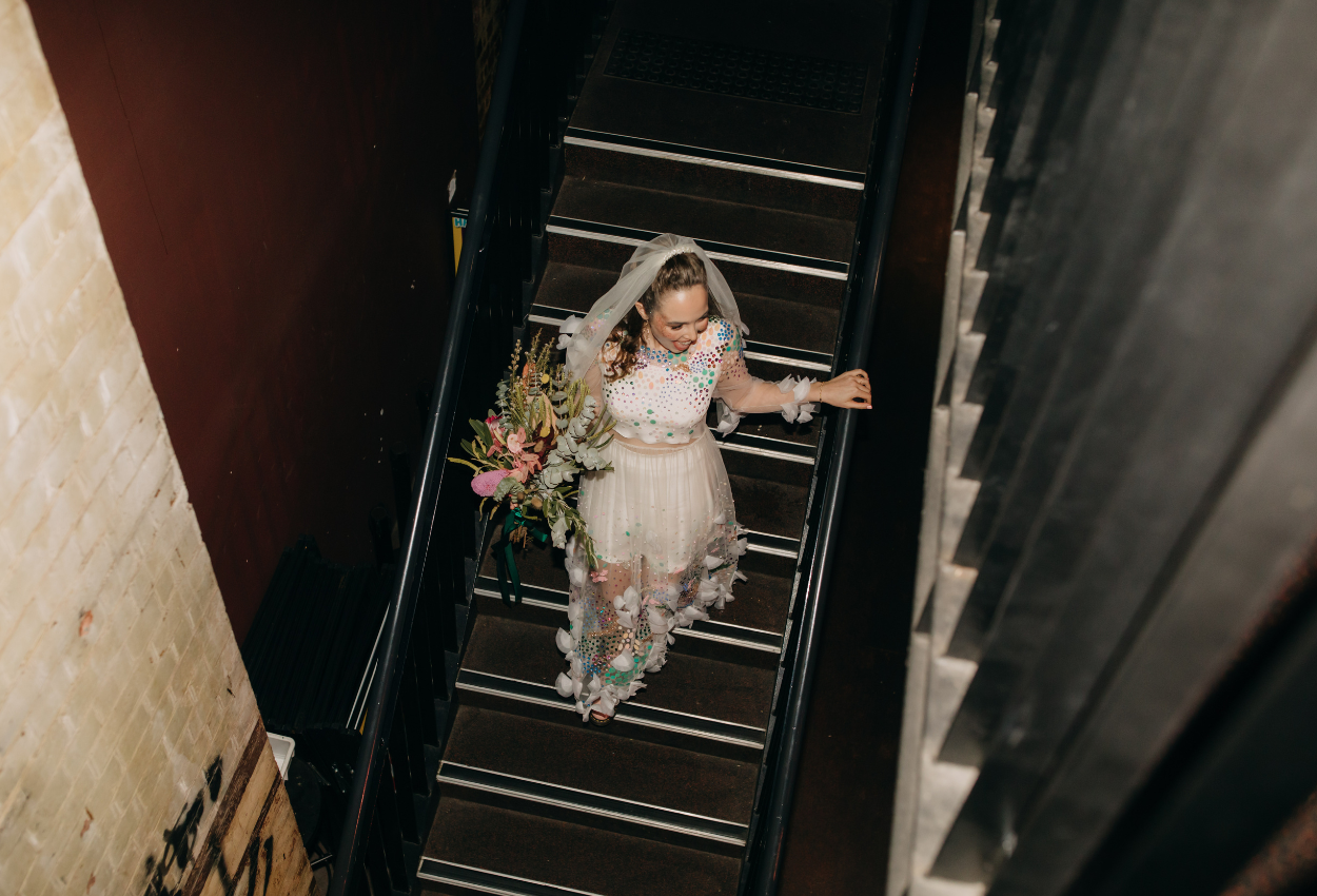 A bride in a floral wedding dress gracefully ascends a dimly lit staircase at Brisbane Powerhouse, clutching a bouquet. Captured from above, the scene highlights the intricate details of her gown and veil, beautifully illuminated by the ambient light.