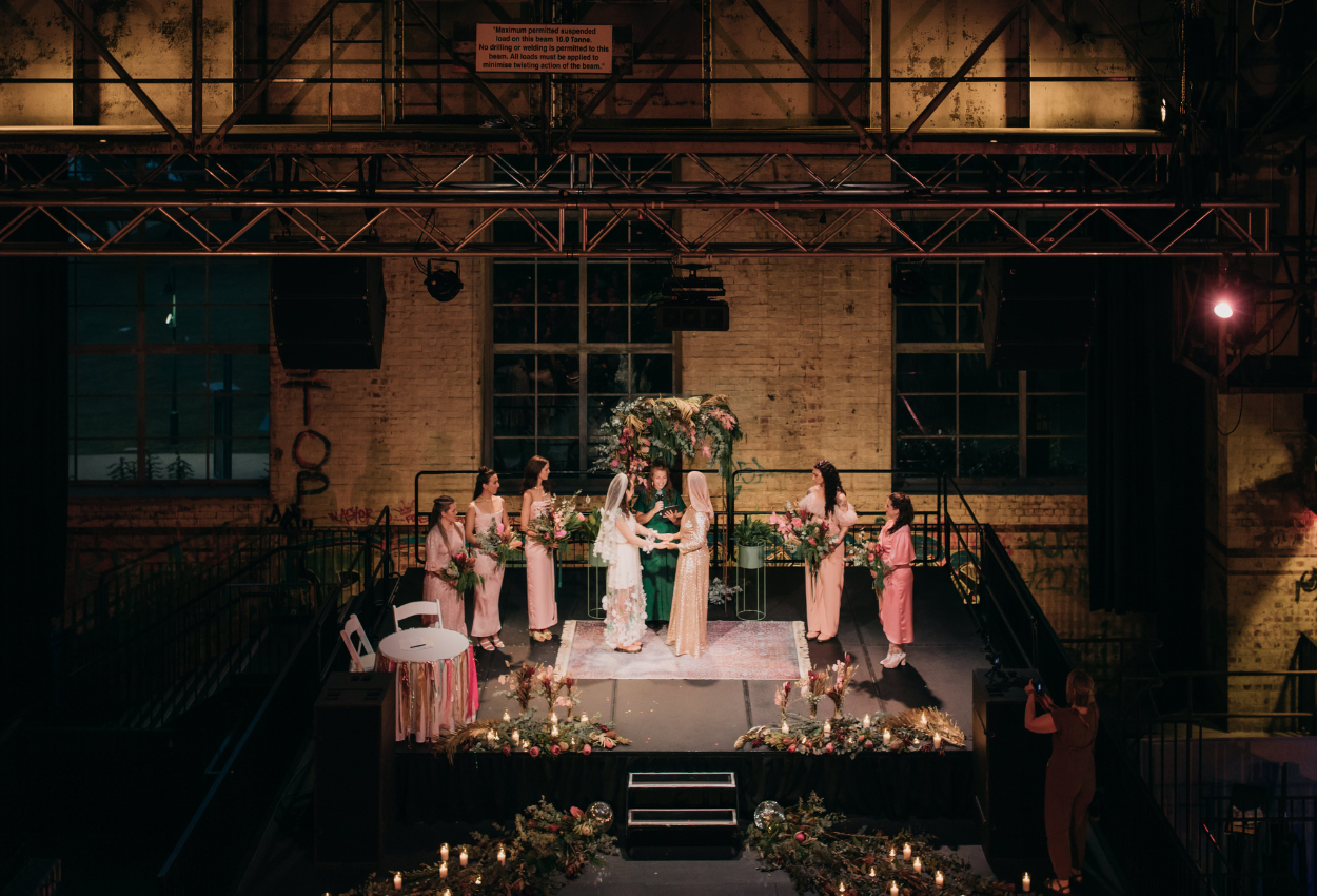 A wedding ceremony in Brisbane Powerhouses industrial-style venue unfolds beautifully. The couple stands on a decorated platform, surrounded by bridesmaids in matching dresses. The area is adorned with flowers and candles, while a photographer captures the moment from below.