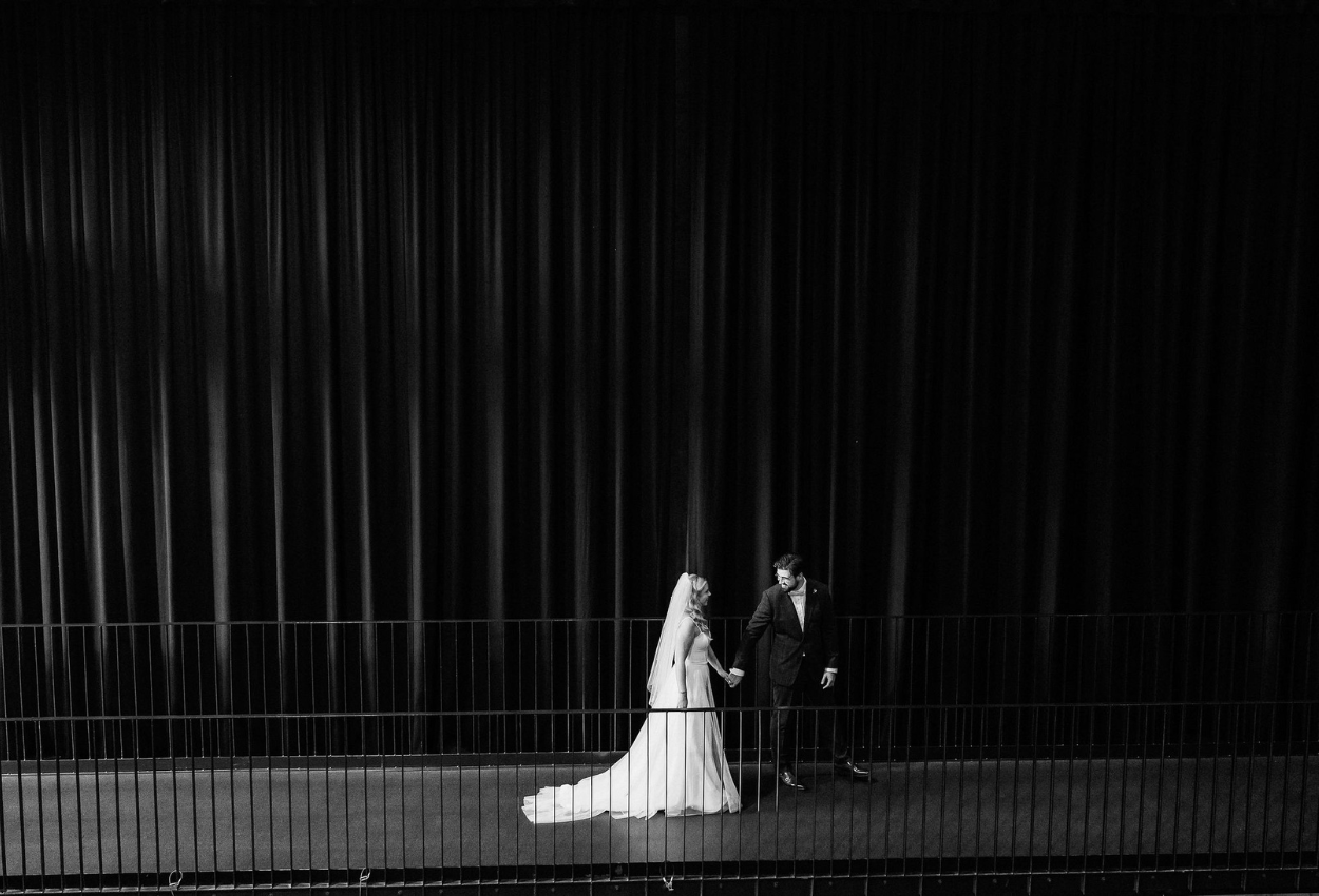 A black and white image of a bride in a long gown and veil, holding hands with a groom in a suit, walking along a corridor at Brisbane Powerhouse. The couple is positioned on the left side of the scene, framed by dark curtains, capturing the elegance of weddings.