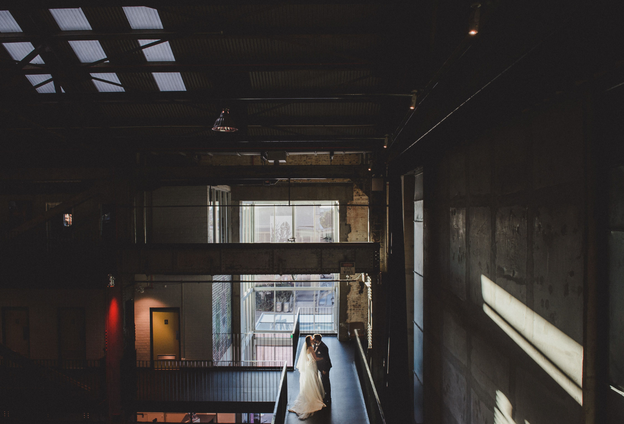 A couple embraces on the modern industrial walkway of the Brisbane Powerhouse, with sunlight streaming through ceiling windows and casting dramatic shadows on the concrete walls. The bride wears a long white gown, beautifully contrasting with the grooms dark suit amidst this unique wedding venue.