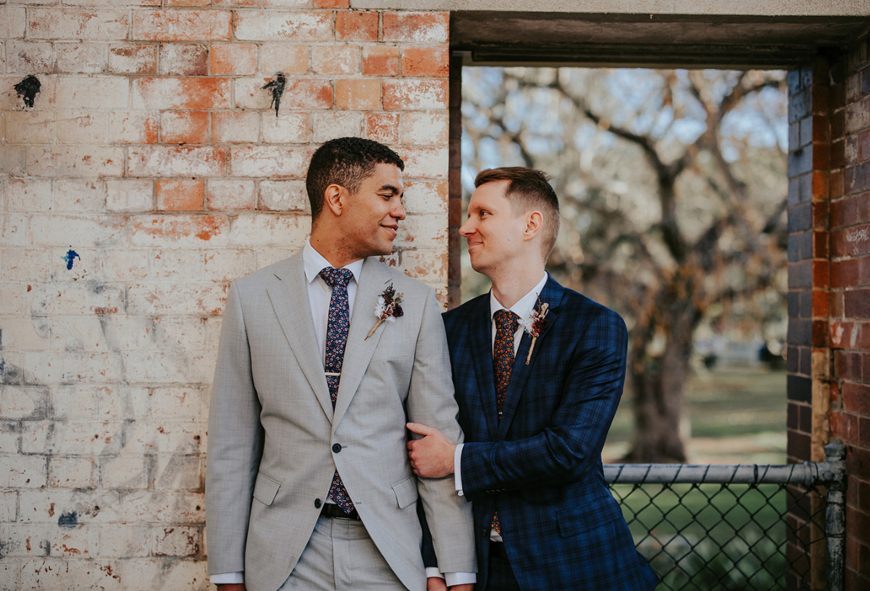 Two people in suits stand closely together, one in a gray suit and the other in a blue plaid suit, exuding joy at what seems to be the perfect setting for weddings. They smile warmly at each other against a rustic brick wall with a window revealing lush trees, at Brisbane Powerhouse.