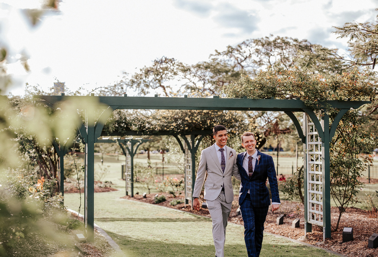 Two people in suits walk under a wooden arbor in a garden, reminiscent of weddings at Brisbane Powerhouse. One wears a gray suit and the other a blue plaid suit. They are smiling and holding hands, surrounded by greenery and flowers on a sunny day.