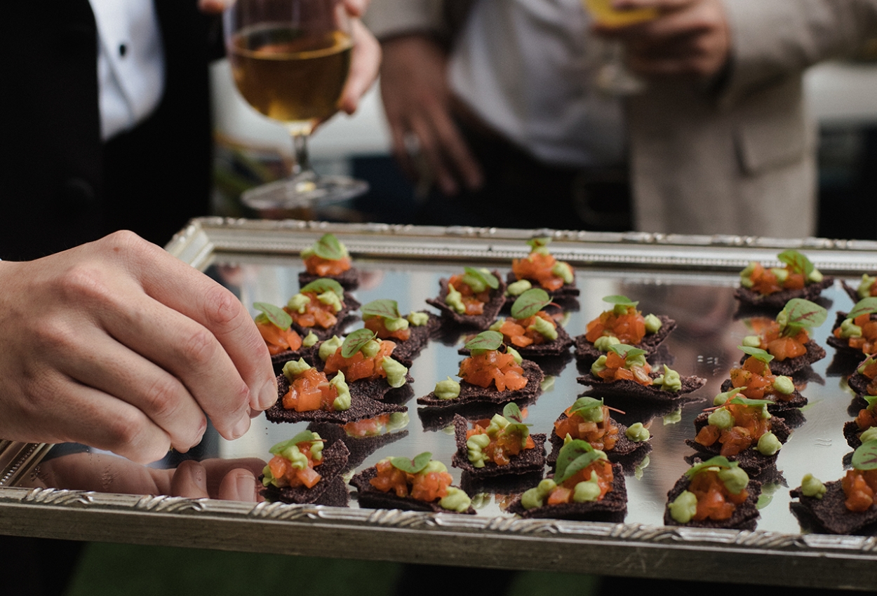 A silver tray holds neatly arranged appetizers topped with diced vegetables and herbs at a stylish social gathering, perhaps a wedding at Brisbane Powerhouse. A hand reaches for one, while someone in the background holds a glass of wine, adding to the lively atmosphere.