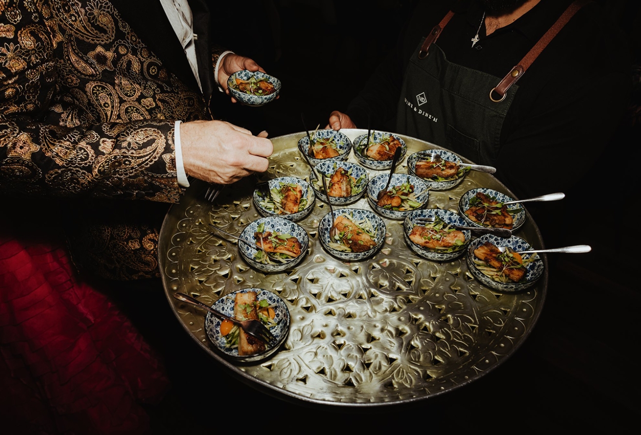 A classy platter with small ornate bowls filled with a colorful salad, likely containing greens, herbs, and sliced vegetables. At a Brisbane Powerhouse wedding reception, a person in a patterned jacket picks one bowl while a server gracefully holds the tray.