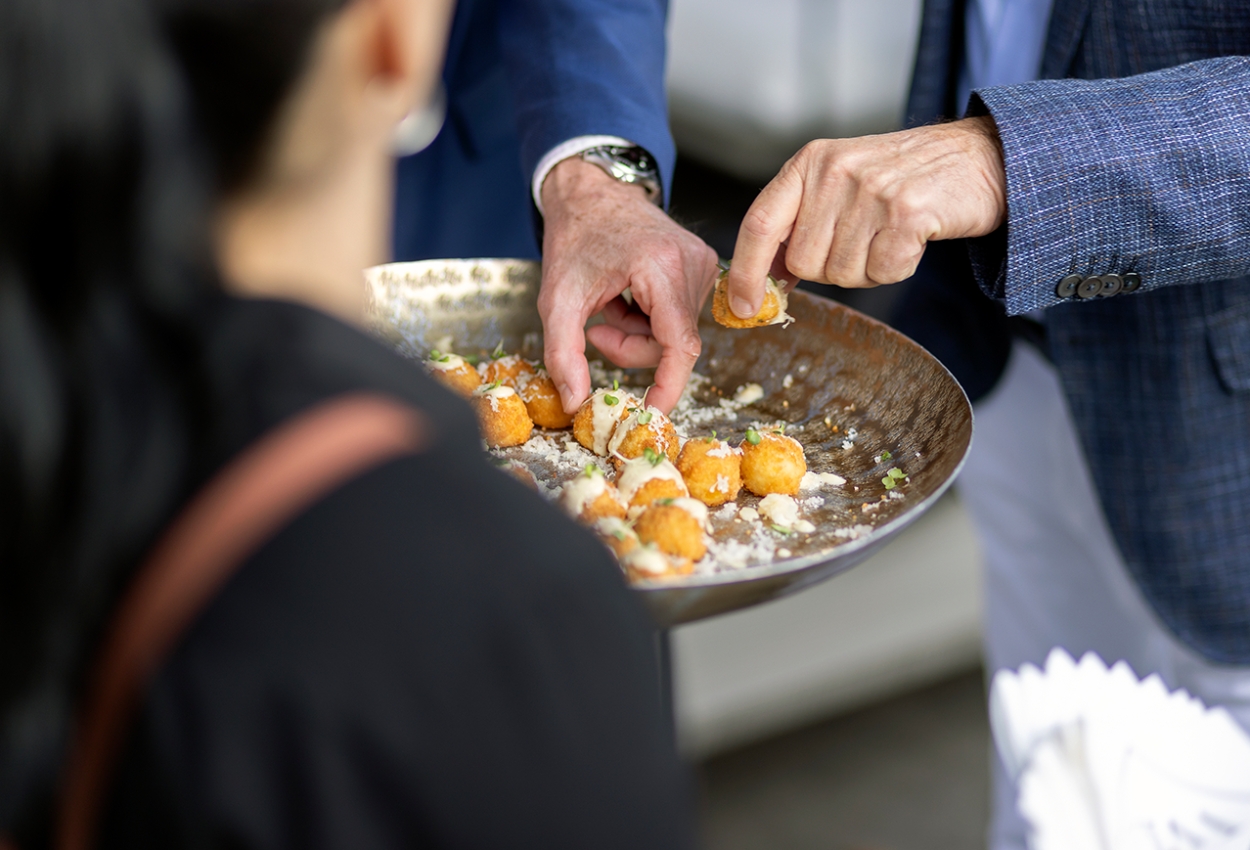 At a sophisticated wedding reception at Brisbane Powerhouse, a person in a suit presents a tray of crispy, bite-sized hors doeuvres, artfully sprinkled with cheese and herbs, to a woman in black.