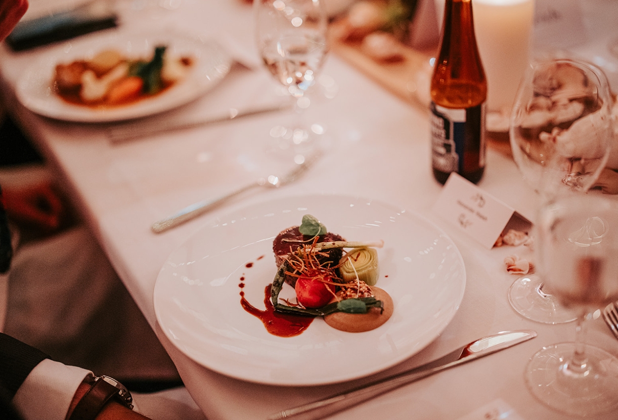 An elegantly plated gourmet dish with colorful vegetables, sauce, and garnish sits on a white plate. The table is set with wine glasses, a beer bottle, and a lit candle, echoing the warm and sophisticated atmosphere perfect for weddings at the Brisbane Powerhouse.