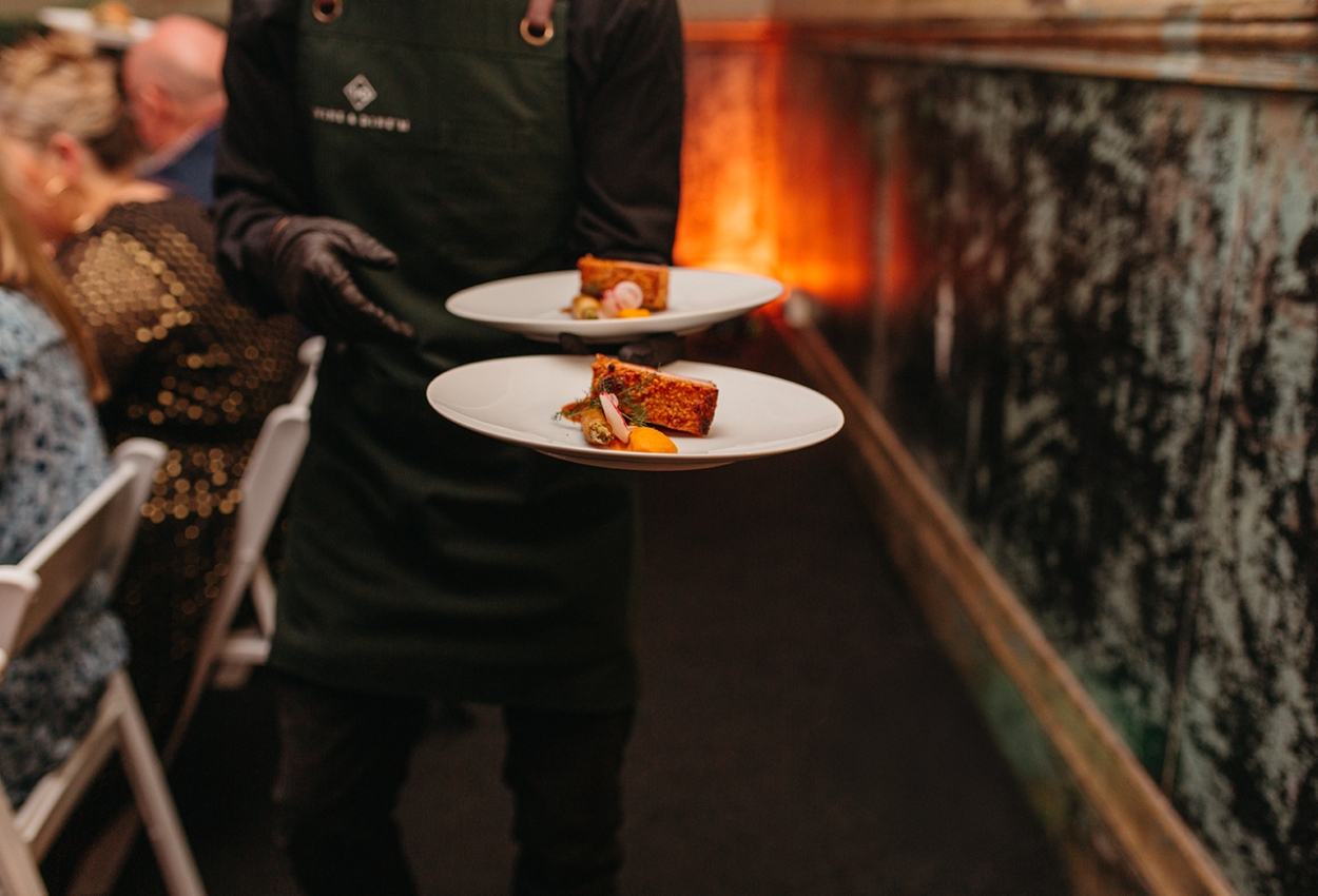 A waiter in a dark uniform serves two plates with gourmet dishes, featuring a meat slice and garnishes. The background captures the elegant ambiance of weddings at Brisbane Powerhouse, with seated guests amidst ornate wall decor and a lively dining scene.