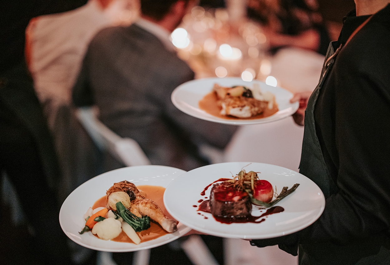 A server gracefully navigates the dimly lit dining area of the Brisbane Powerhouse during a wedding reception, carrying three plates of elegant gourmet dishes. In the background, blurred diners enjoy artfully arranged meals featuring vibrant vegetables and exquisite sauces.