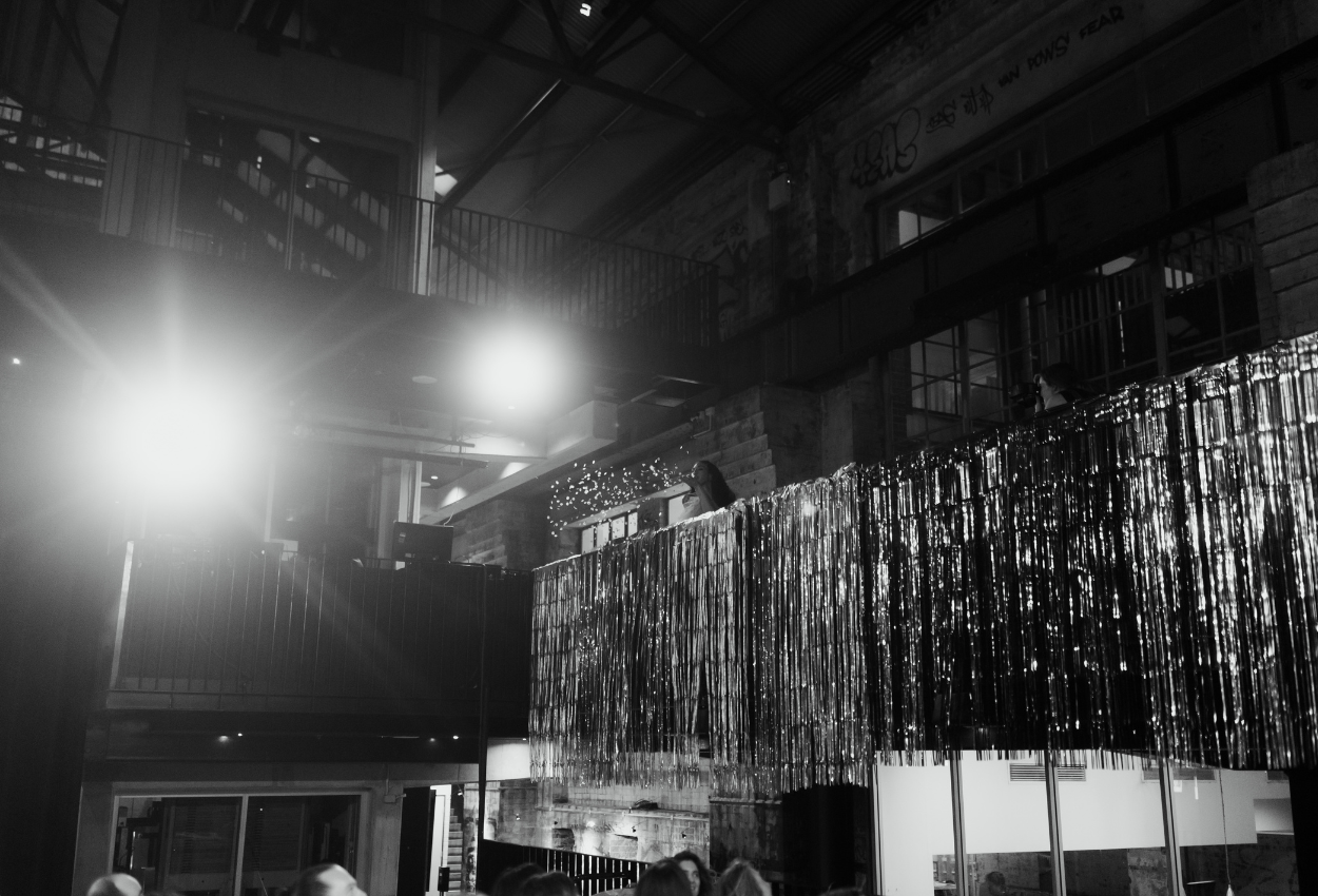 A black and white image of the Brisbane Powerhouses industrial interior with high ceilings and exposed brick captures a unique wedding scene. Bright lights illuminate tinsel curtains along a balcony, while guests gather below and decorative lights twinkle on the mezzanine.