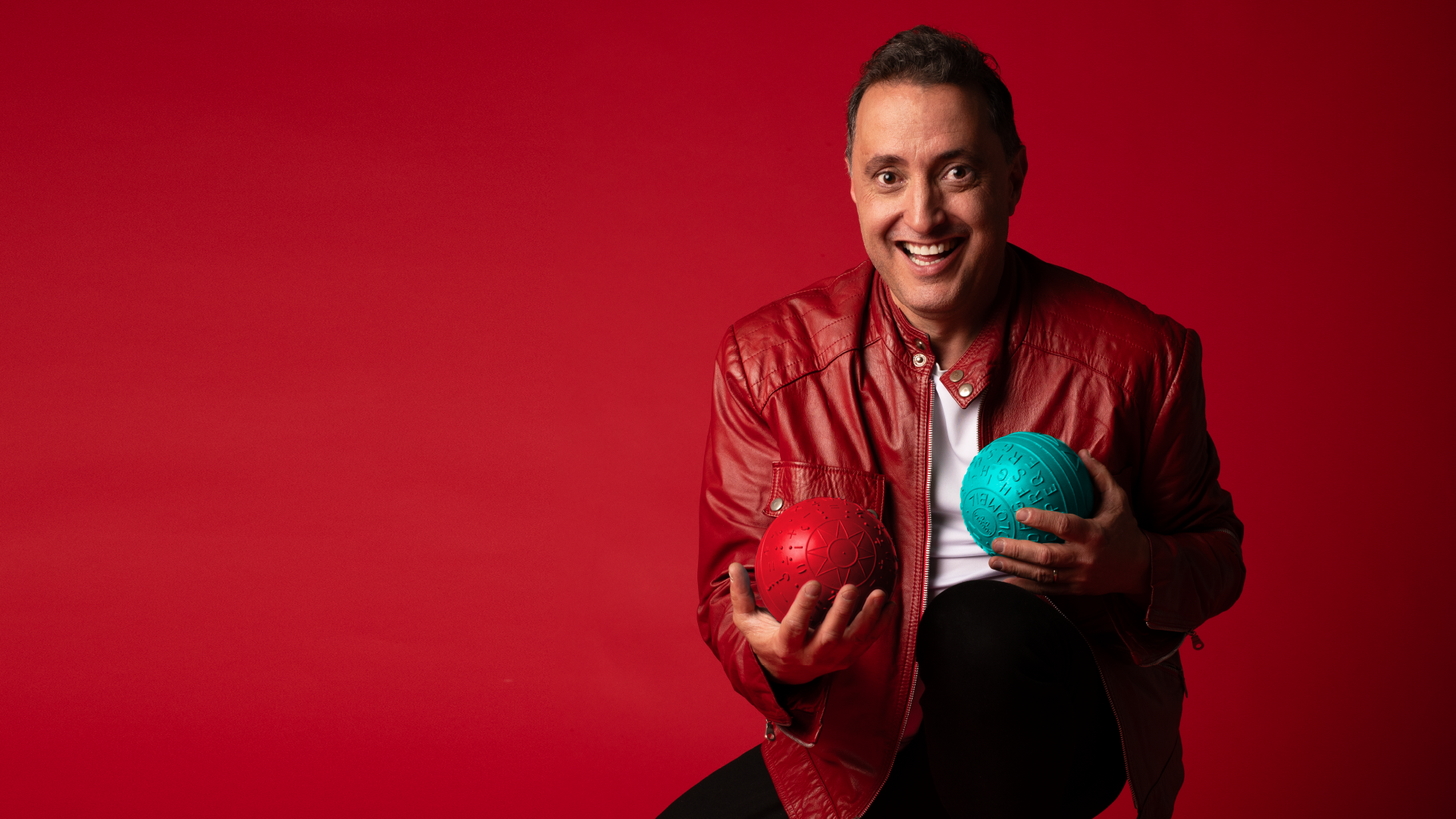 Andres Lopez, dressed in a striking red leather jacket, smiles while holding a vibrant red and blue ball against a bold red background. Catch him at the Brisbane Powerhouse 2025 for an unforgettable experience.