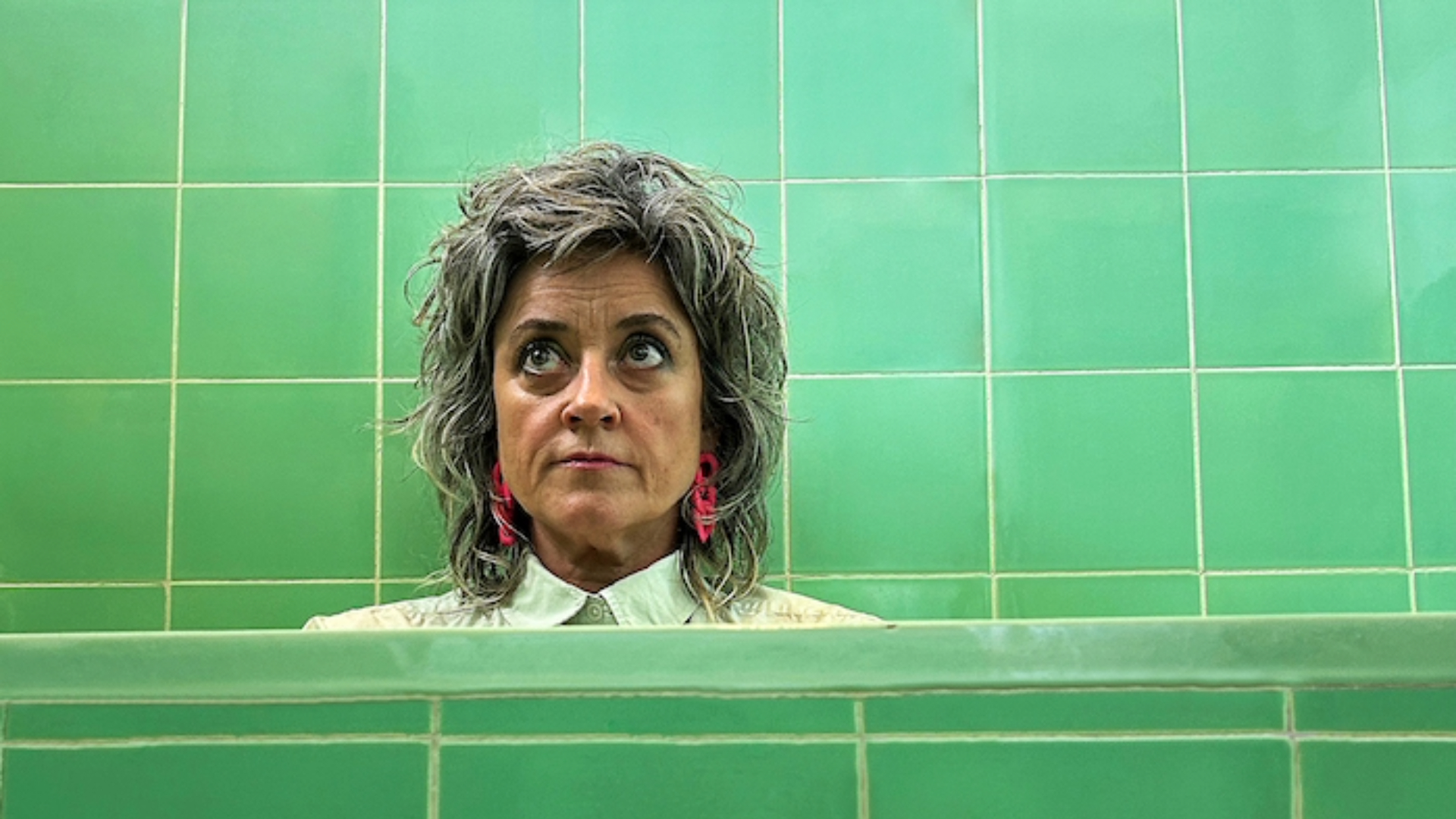 A person with short, tousled hair and pink earrings is standing in front of a green tile wall, looking up with an expression of contemplation or curiosity.