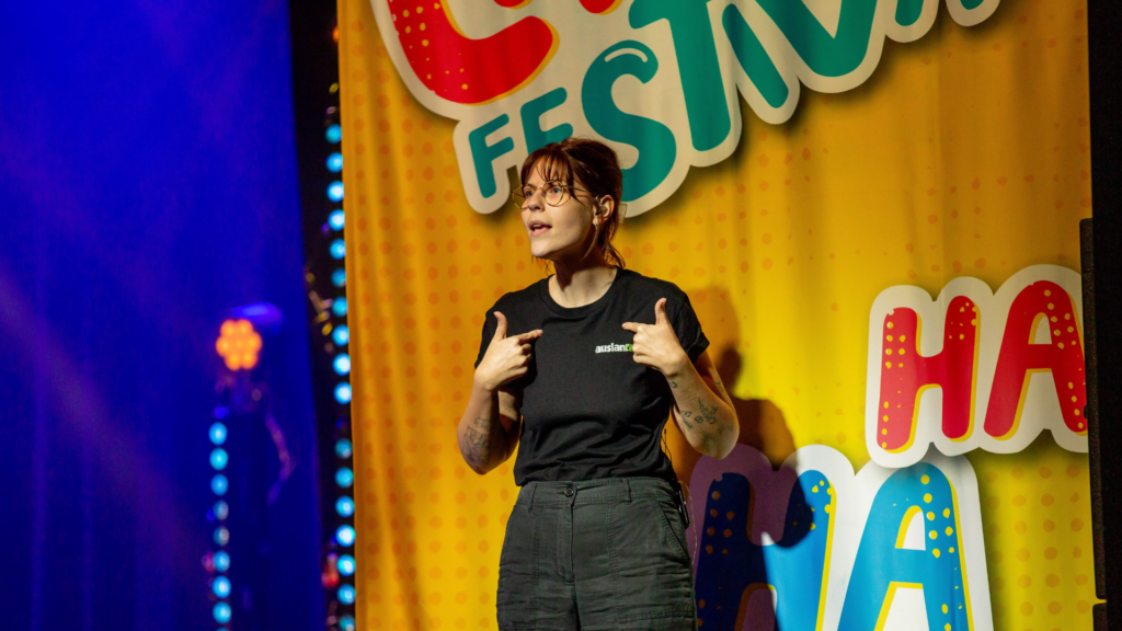 A person stands on stage pointing at themselves and speaking animatedly. They wear glasses and a black shirt. Behind them is a colorful banner with festive text and patterns. Stage lights create a vibrant atmosphere.