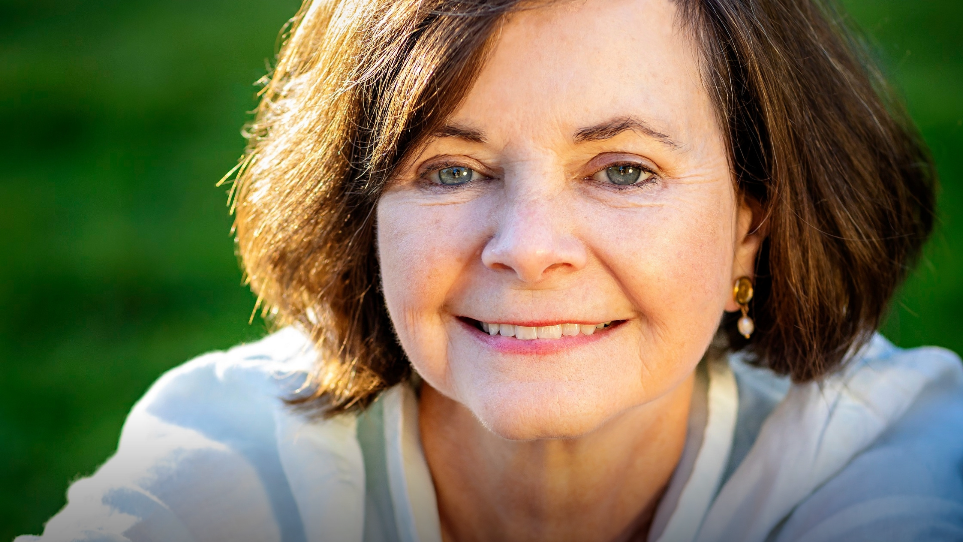 A woman with short brown hair smiles warmly, sitting outdoors on a sunny day, reminiscent of a scene from a Geraldine Brooks novel. Her white top and earrings glisten under the sun, while the blurred green backdrop conjures thoughts of an inviting Brisbane Powerhouse garden in 2025.