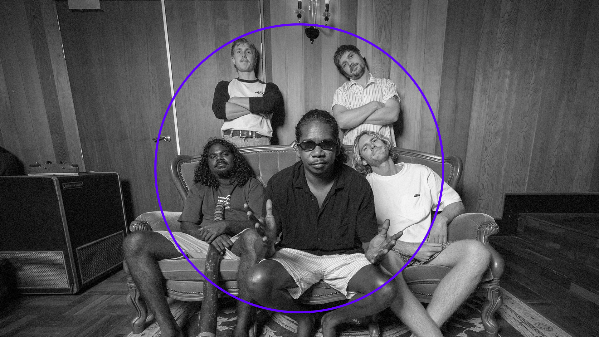 A black-and-white photo captures the spirit of the OHM Festival 2025. Five people gather in a room, with two seated on a couch, one gesturing animatedly. Three others stand behind them, encircled by a purple outline. Wooden walls and a hanging light frame this evocative moment.