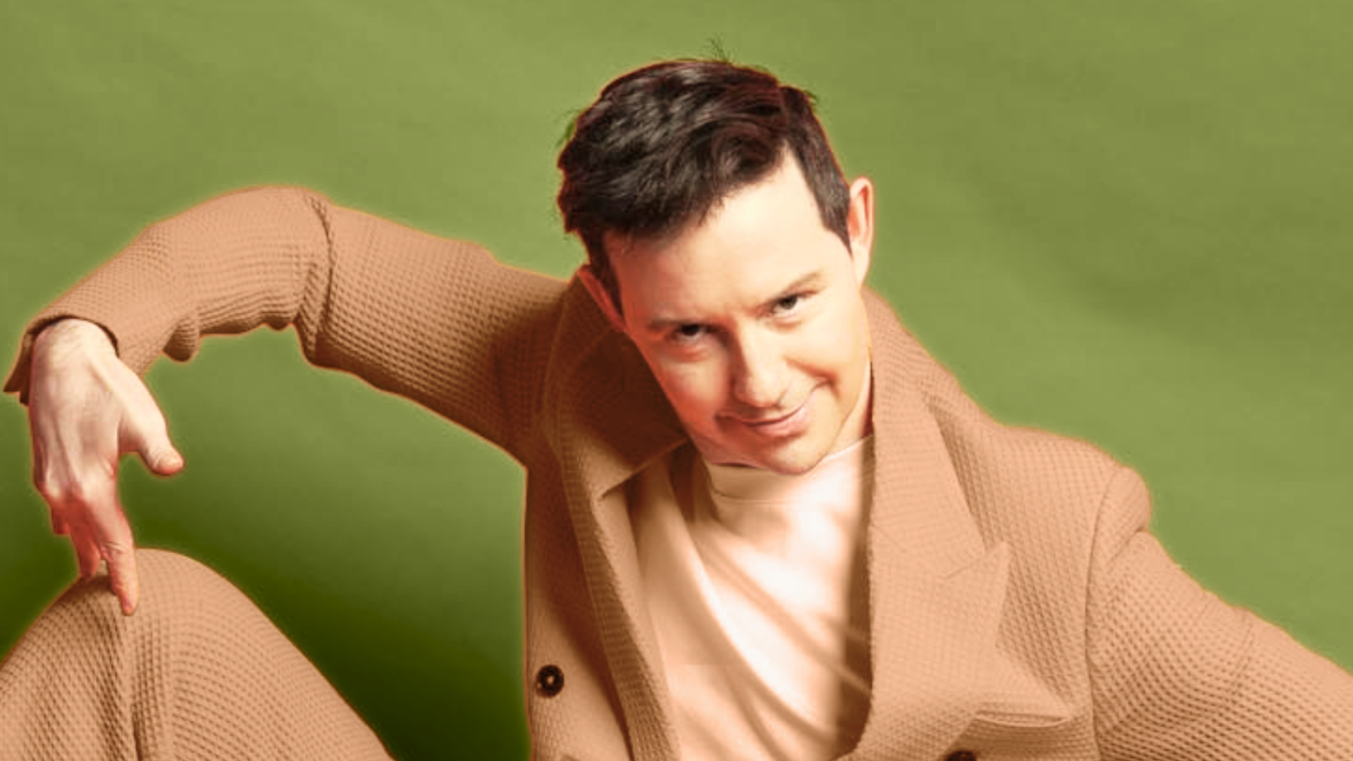 A person with short dark hair wearing a beige textured suit and a white shirt poses against a green background, smiling at the camera. Their right arm is bent upwards and their left knee is raised.