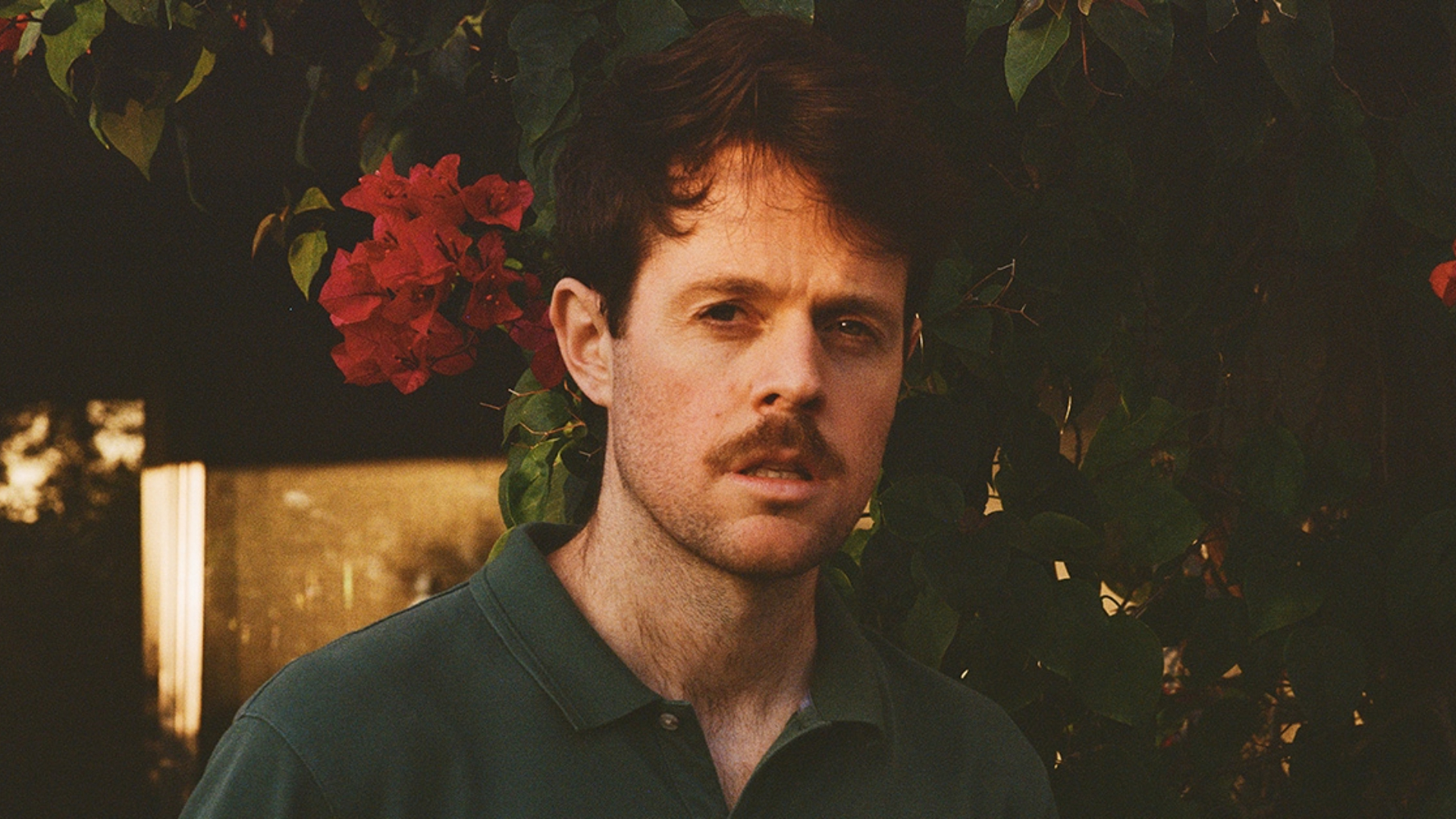 A person with short, dark hair and a mustache stands outdoors in front of greenery with red flowers. They are wearing a dark green collared shirt, and the background is softly lit by natural light.