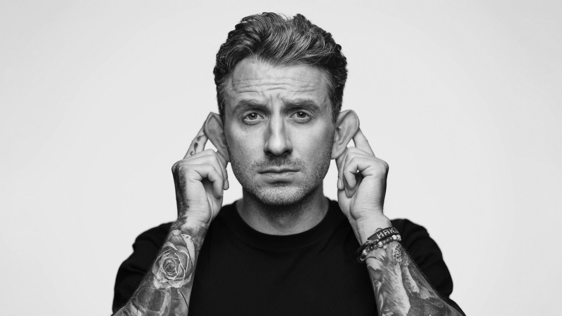 A person with tattoos on their arms playfully pulls their ears to the side, making an exaggerated ear gesture. They are wearing a black t-shirt and looking directly at the camera against a plain background.