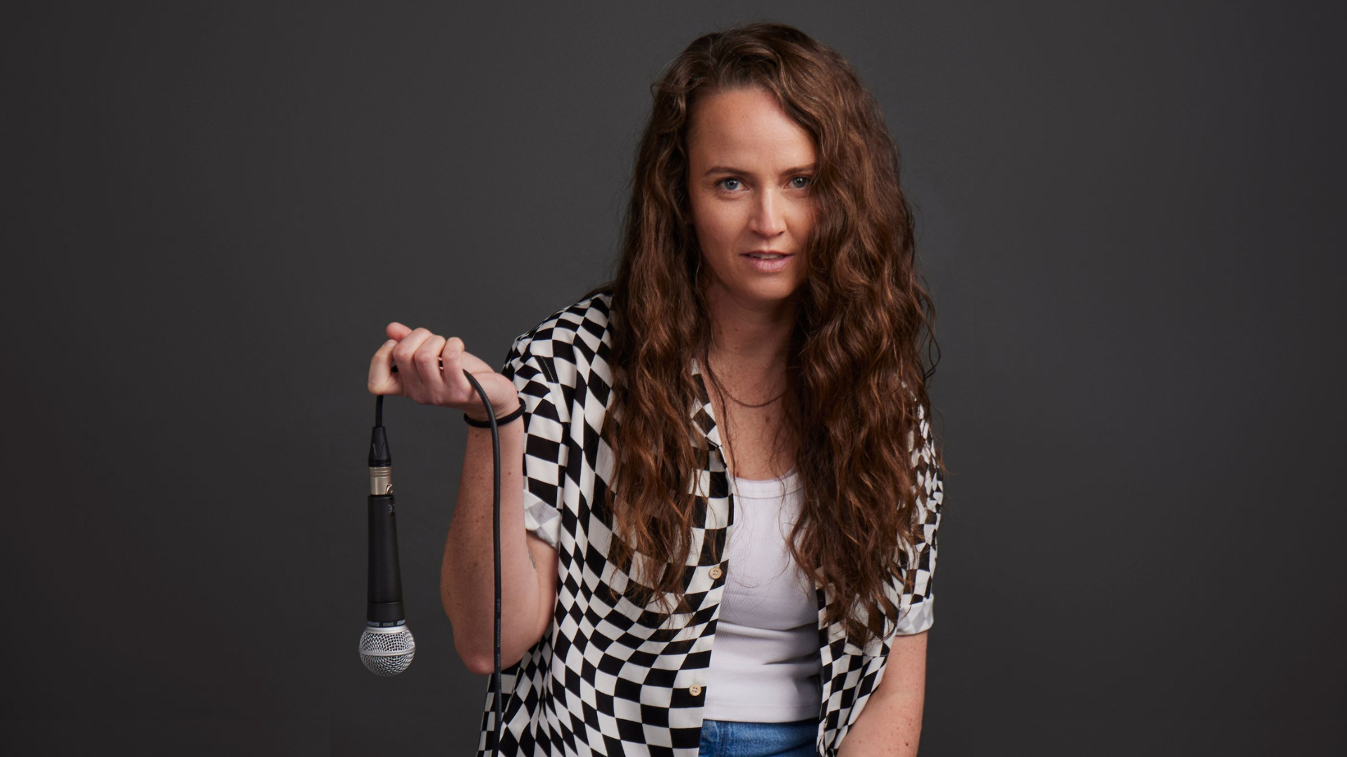 A woman with long curly hair holds a microphone casually. She wears a black and white checkered shirt over a white top and jeans, posing against a dark background.