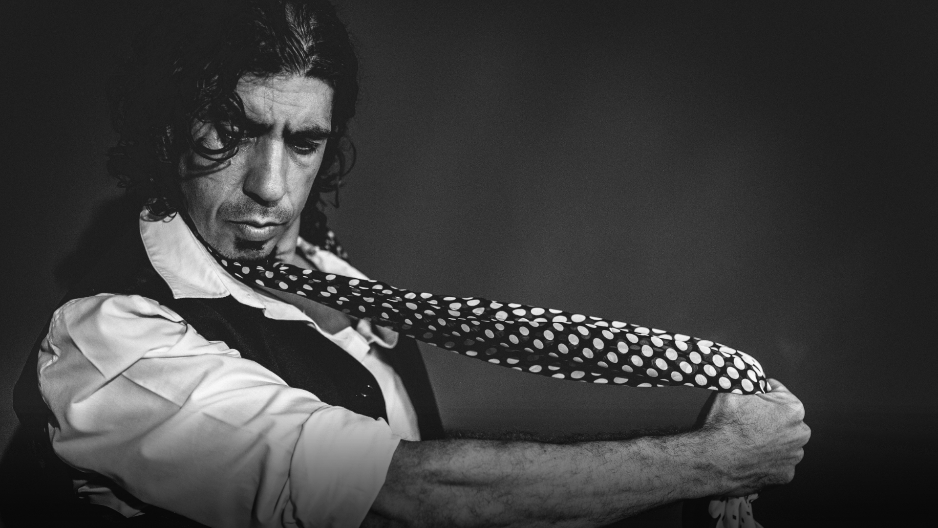 A man with long, curly hair is intensely looking downwards as he adjusts a polka dot scarf around his neck. Reminiscent of the passion in *Poemas de Iberia*—coming to Brisbane Powerhouse 2025—he wears a white shirt and dark vest against a shadowy backdrop.