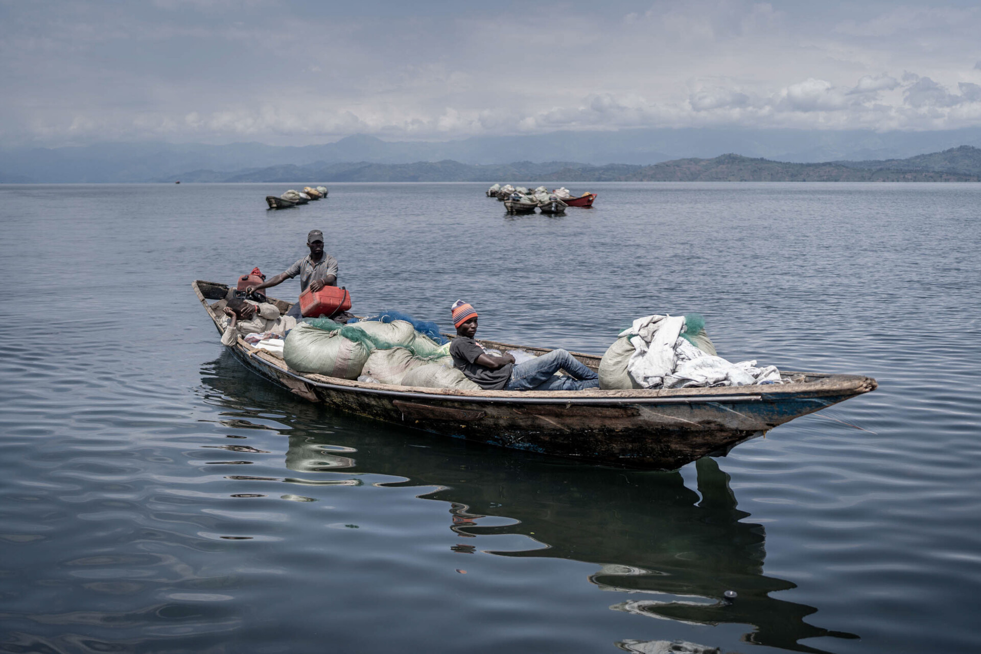 Three men sit in a wooden boat filled with sacks and bags, floating on calm water with distant hills and cloudy skies behind. This evocative scene could be featured at the World Press Photo Exhibition 2025 at Brisbane Powerhouse.