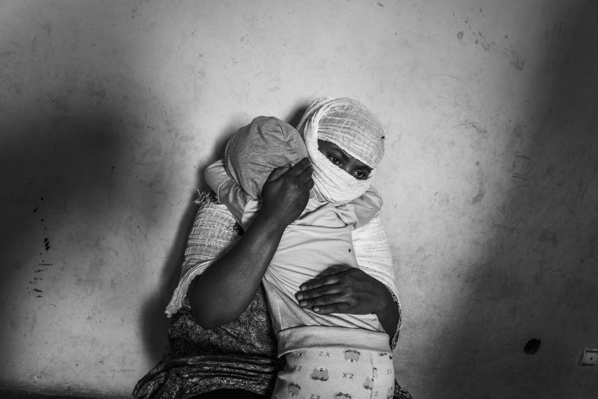 A woman with bandaged arms and head sits against a bare wall, comforting a child whose face is covered with a shirt—a powerful scene of vulnerability featured in the World Press Photo Exhibition 2025 at Brisbane Powerhouse.