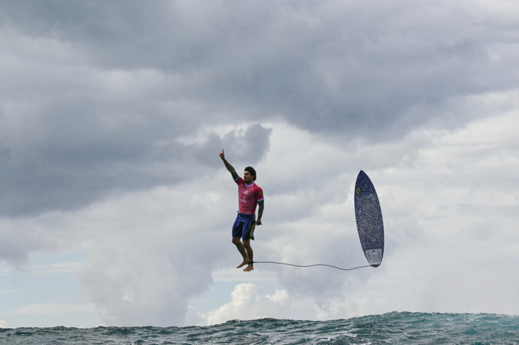 A surfer in a pink shirt and blue shorts is airborne above the ocean, separated from his surfboard, with one arm raised and dramatic clouds in the sky behind him.