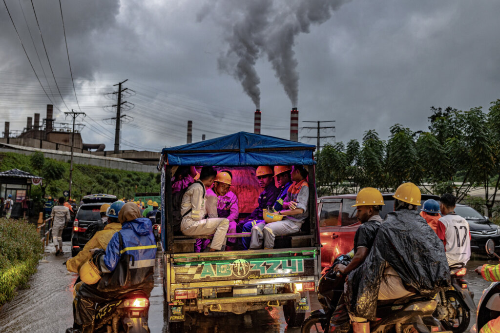 A group of workers in safety gear sit in the back of a truck with purple lighting, surrounded by motorbikes and cars, while factory smokestacks emit thick smoke under a cloudy sky in the background.