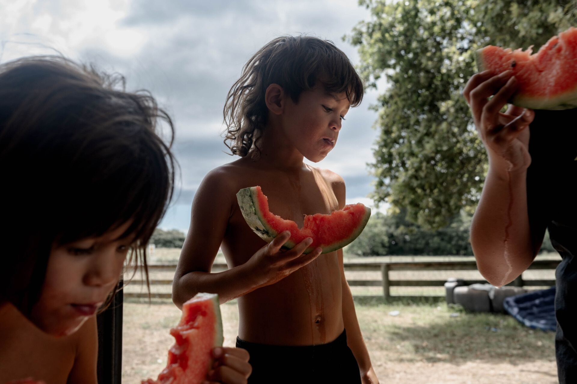 Three children stand outside eating slices of watermelon. One child at the center looks down, holding a large slice, while another and an adult, both partially visible, eat theirs. Trees and a fence are in the background at the World Press Photo Exhibition 2025 Brisbane Powerhouse.