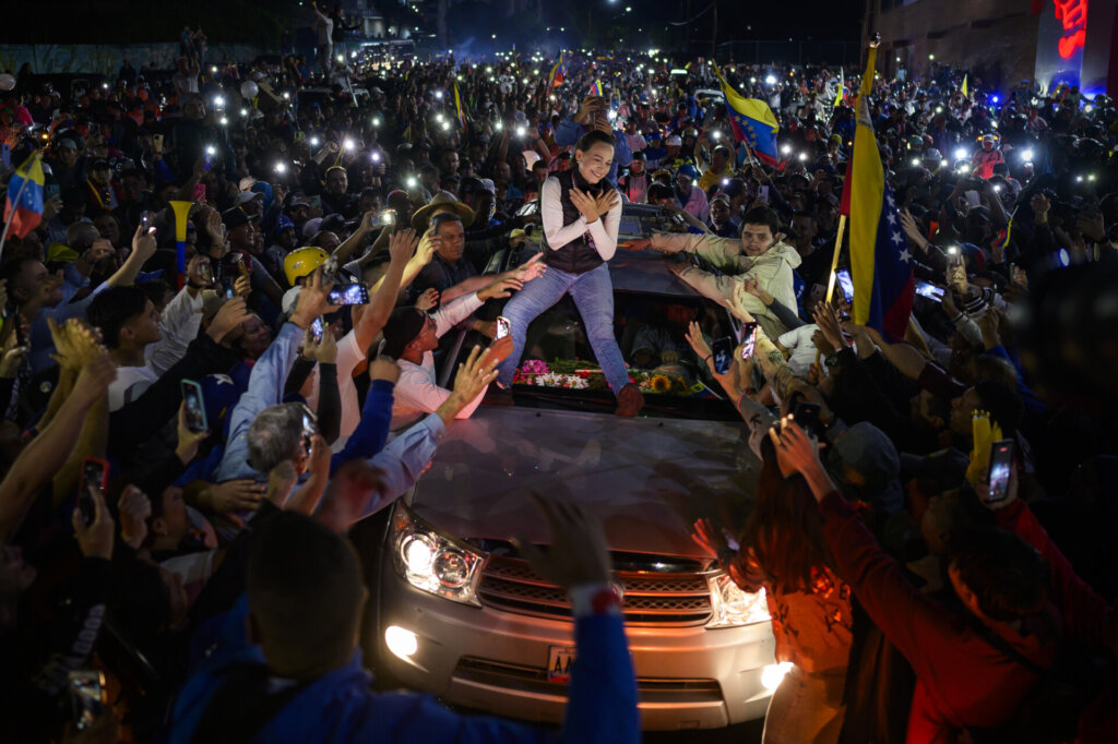 A woman stands on the hood of a car, surrounded by a crowd of people taking photos and reaching toward her, while Venezuelan flags are visible and people hold up their phones in a nighttime scene.