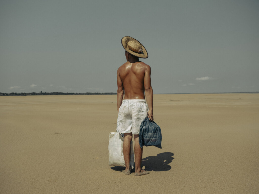 A shirtless person wearing a straw hat and white shorts stands on a vast sandy beach, holding a white bag and a blue bag, looking toward the horizon under a clear sky—a scene reminiscent of the World Press Photo Exhibition 2025 Brisbane Powerhouse.