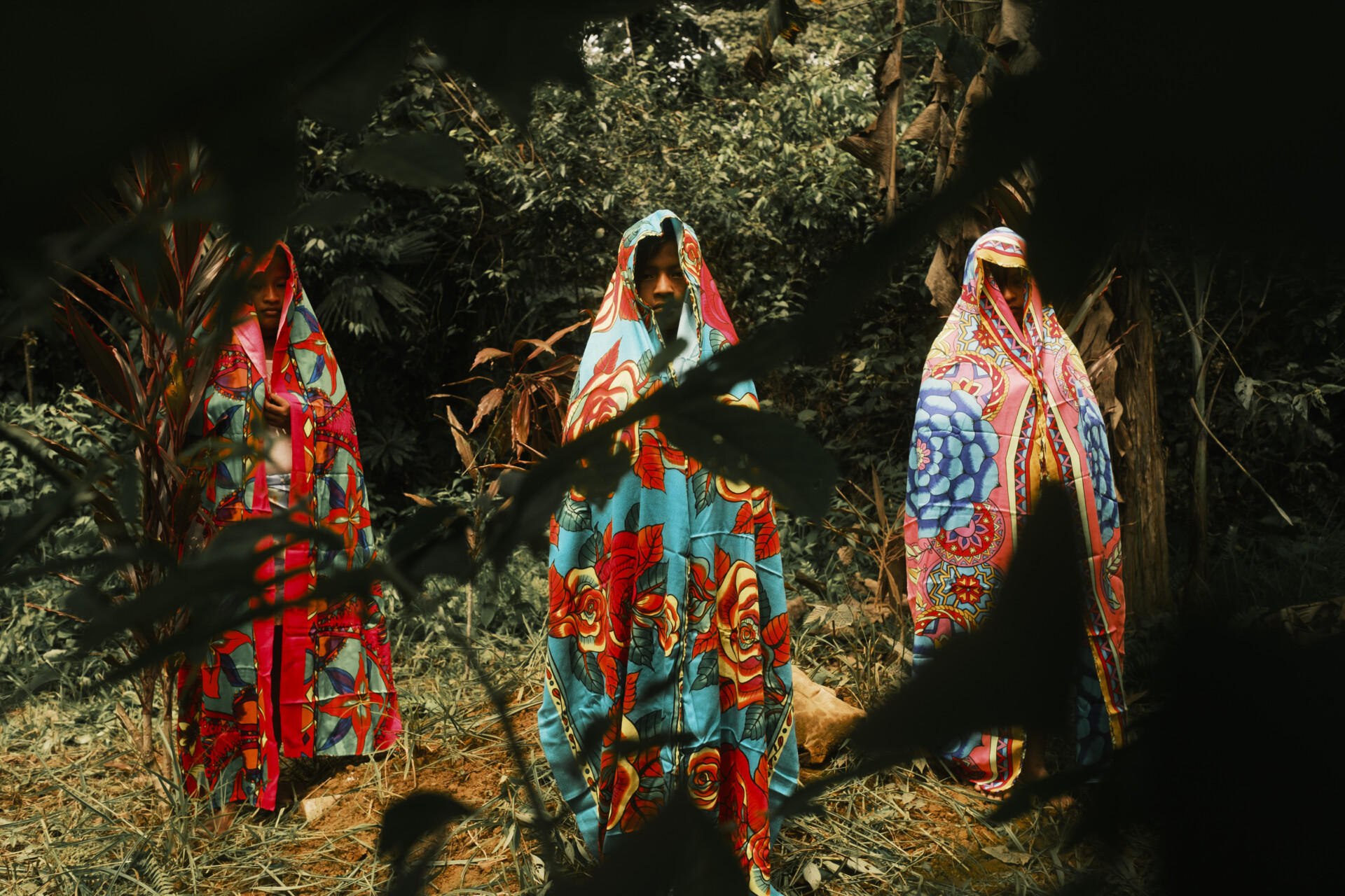 At the World Press Photo Exhibition 2025 Brisbane Powerhouse, three people stand in a forest, each covered head-to-toe in vividly patterned, colorful fabrics, with only their faces visible. The scene is partly obscured by dark leaves in the foreground.