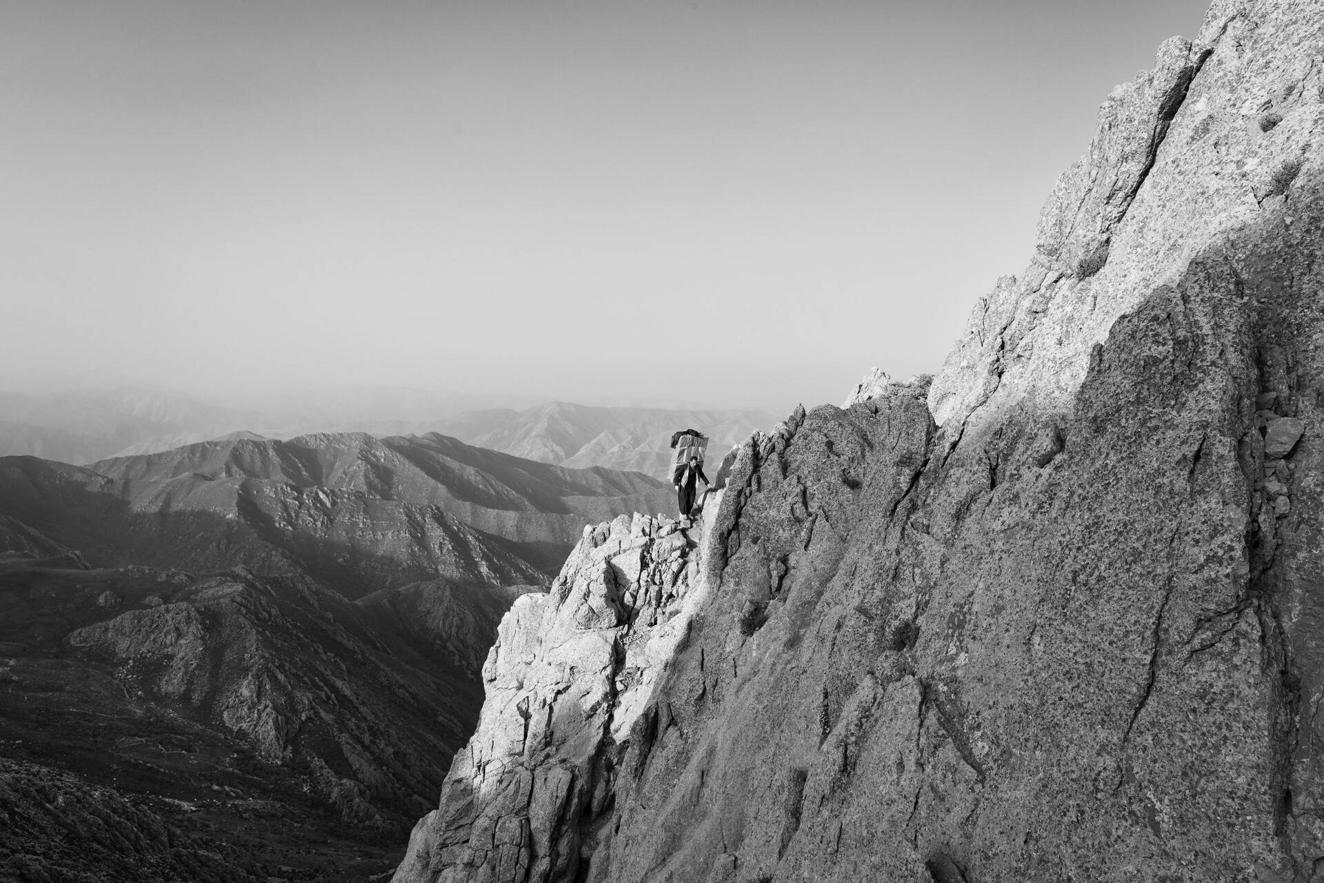 A person with a backpack climbs a steep rocky mountain face, surrounded by rugged peaks and valleys under a clear sky. In black and white, the dramatic solitude echoes images from the World Press Photo Exhibition 2025 Brisbane Powerhouse.