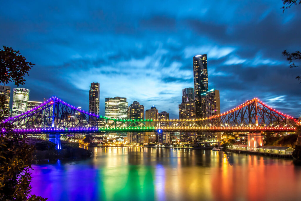 A city skyline at dusk with a brightly lit bridge displaying rainbow colors over a river, reflecting vibrant lights on the water.