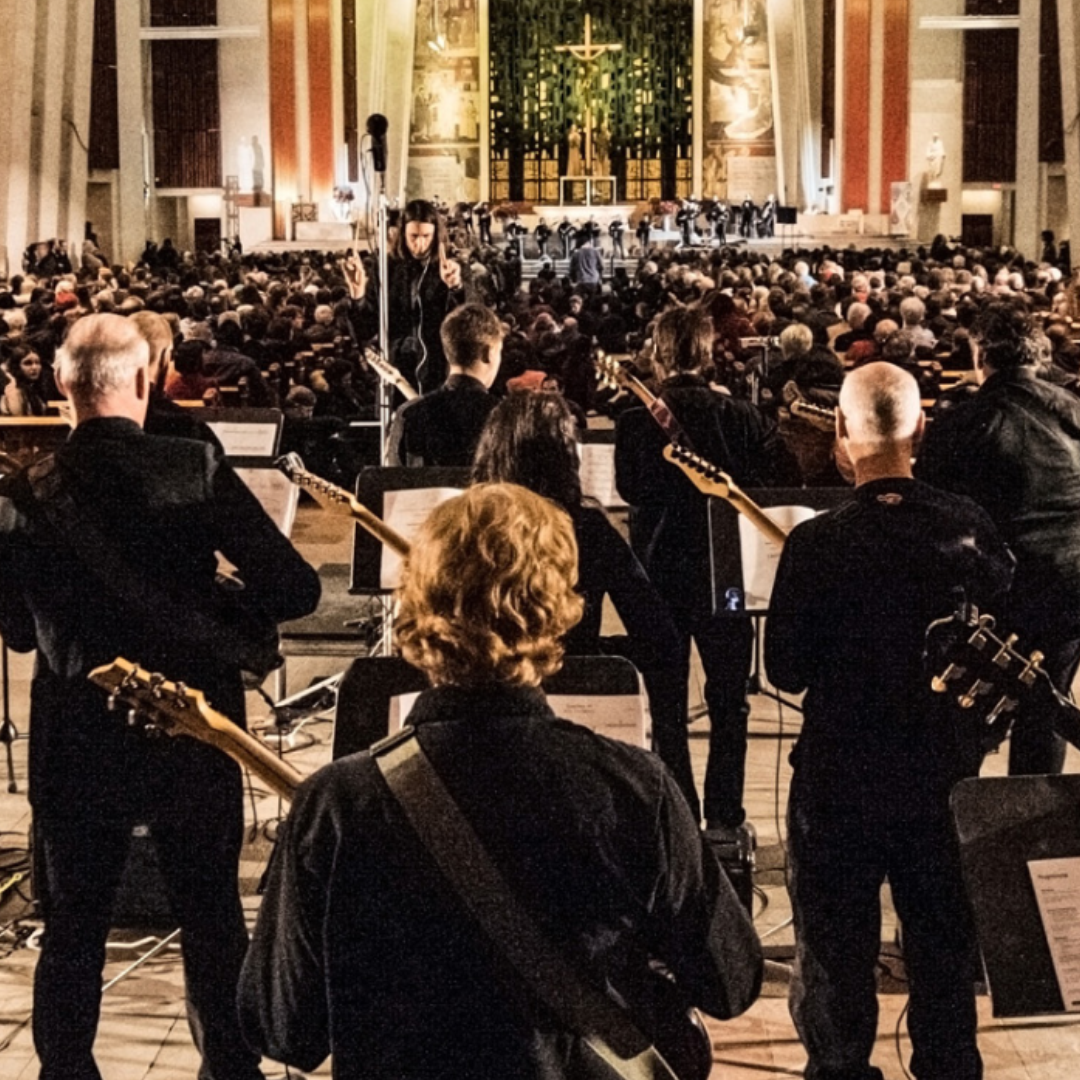 A large orchestra and choir perform in a grand, well-lit church. Musicians with instruments and sheet music face the conductor and altar before a seated audience.