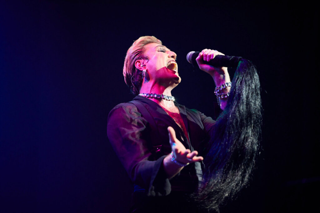 A singer with short blond hair passionately performs on stage, holding a microphone with a dramatic black fringe. The singer wears a black outfit, silver jewelry, and is illuminated by stage lights against a dark background.