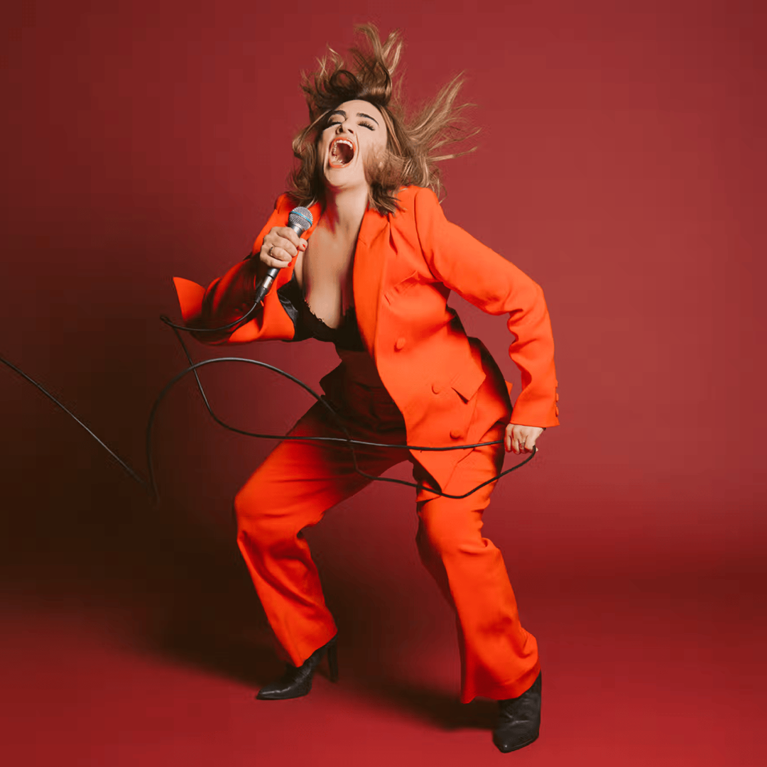 A woman in an orange suit passionately sings into a microphone, with her hair flying and microphone cable swirling, against a red background.