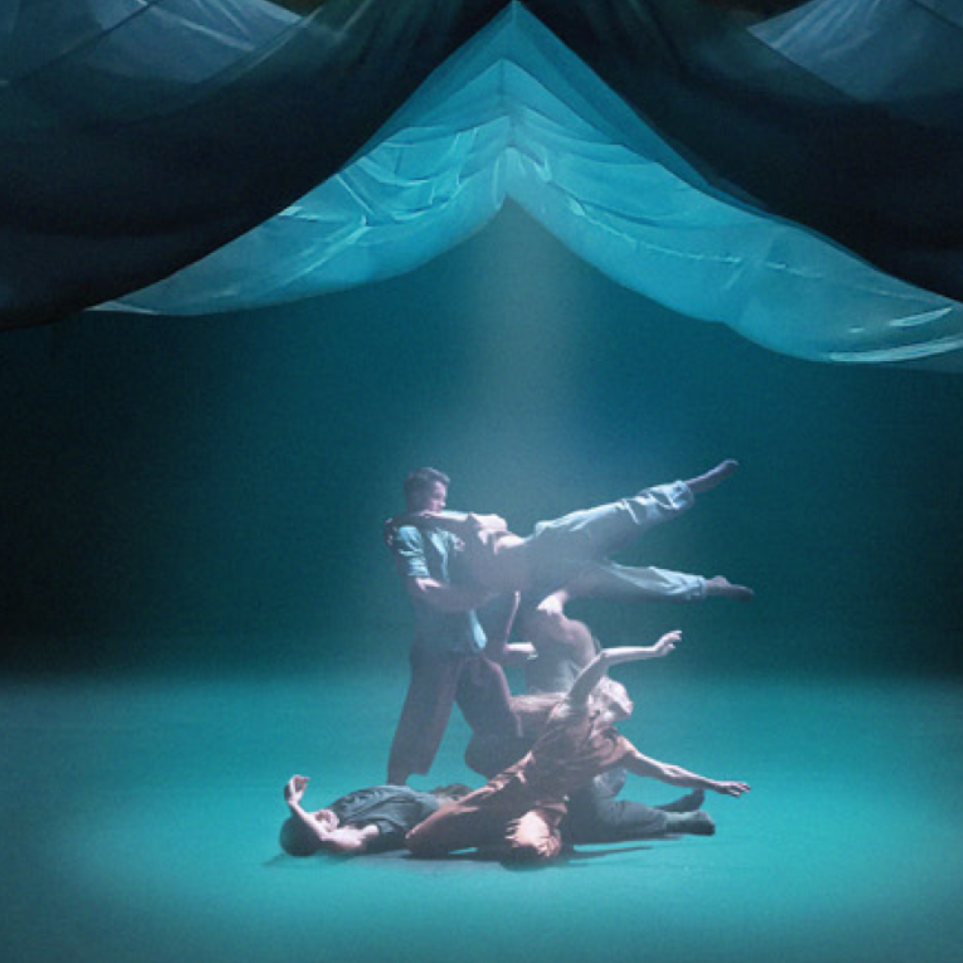 Four dancers perform on a dimly lit stage in Bad Nature with one lifted horizontally by two others beneath billowing blue fabric that hangs overhead, creating a dramatic and ethereal atmosphere.