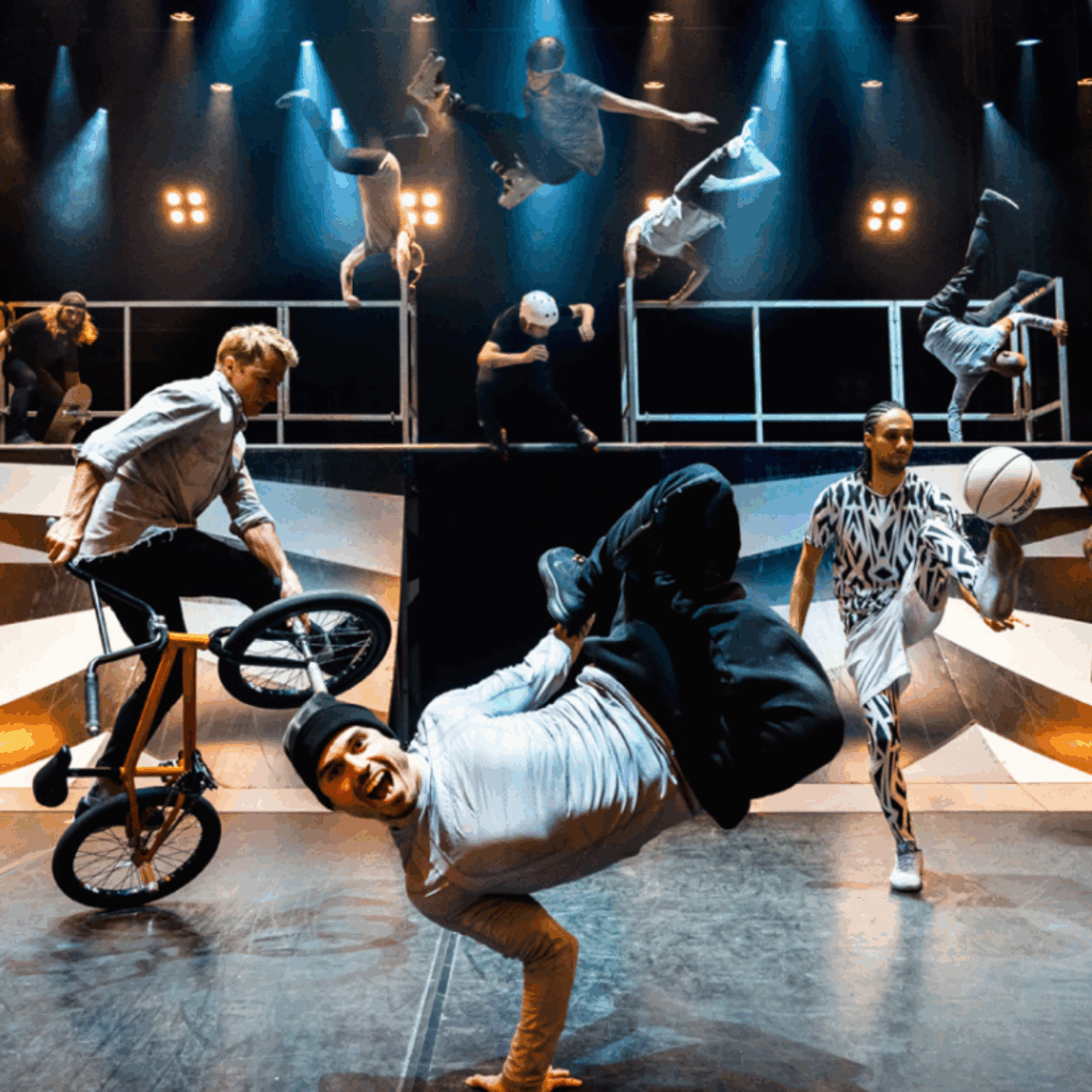 A group of performers breakdancing, skateboarding, and doing BMX tricks on stage under bright spotlights at Elements of Freestyle showcasing energetic street and acrobatic skills.