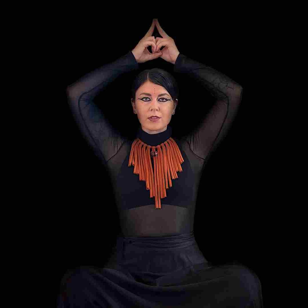 A person in black sheer clothing poses against a black background, arms raised above their head with fingers touching to form a triangle. They wear a bold orange necklace and dramatic eye makeup, evoking the avant-garde spirit of IRTYU at Brisbane Powerhouse.