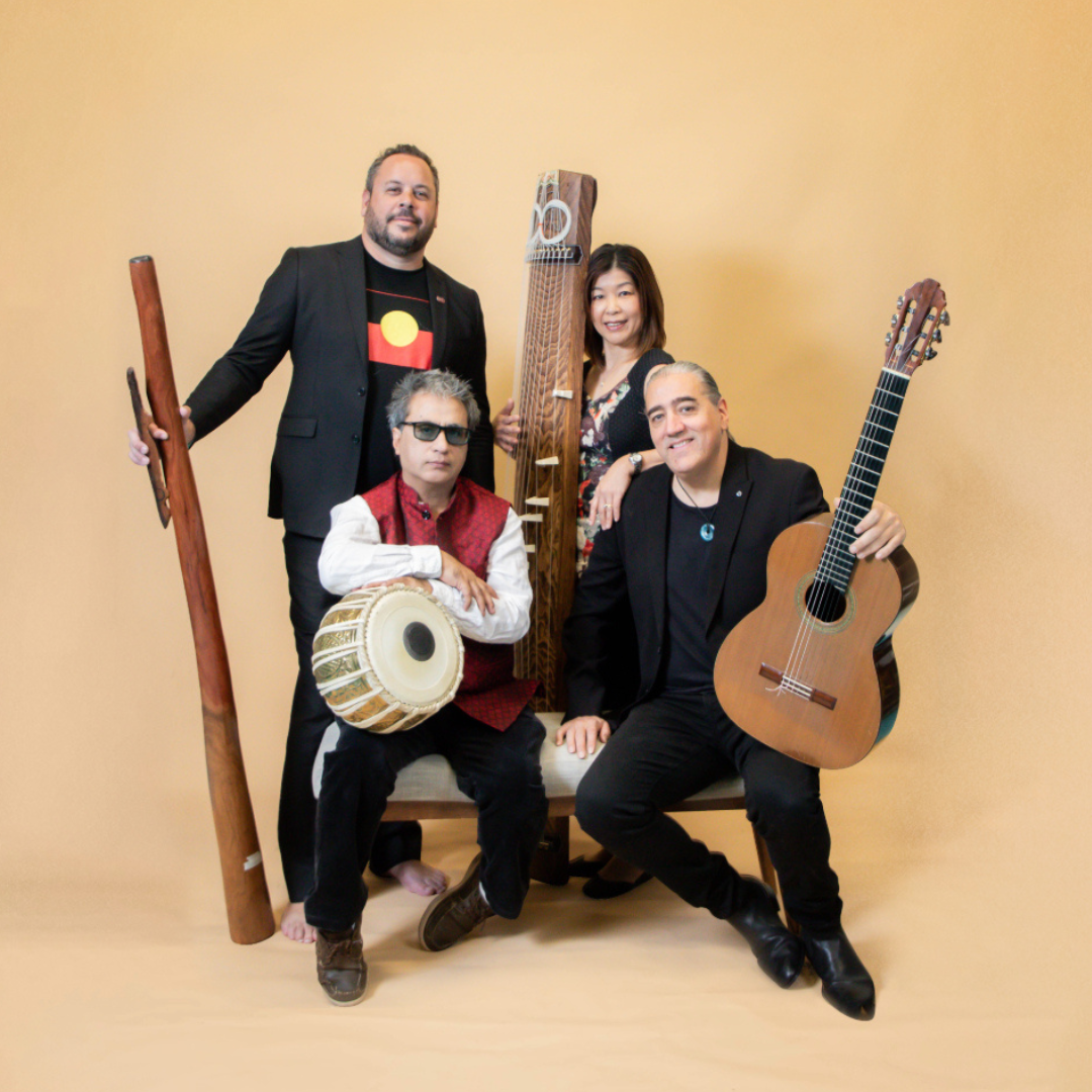 Four musicians pose together against a beige backdrop, holding a didgeridoo, tabla, koto, and guitar. Two are seated while two stand behind, all smiling or looking content with their instruments.
