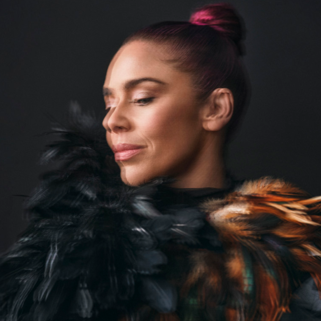 A woman with pink hair in a bun looks down, wearing an elaborate black and orange feathered garment against a dark background.