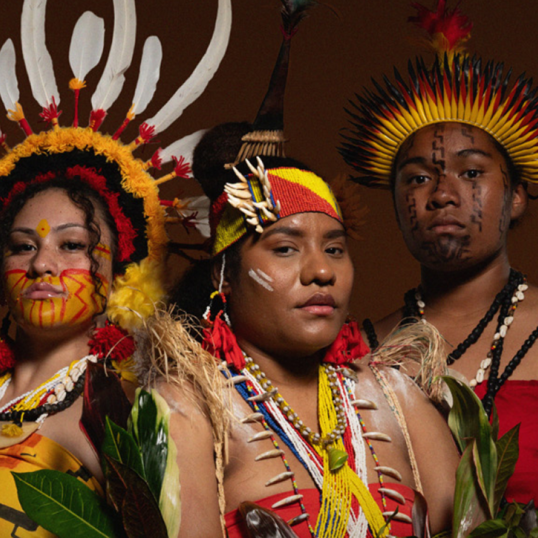Three Indigenous people wear traditional, colorful clothing, feathered headdresses, face paint, and bead jewelry. Standing close together against a dark brown background, they hold green leaves—part of the Pasifika Made at Brisbane Powerhouse celebration.