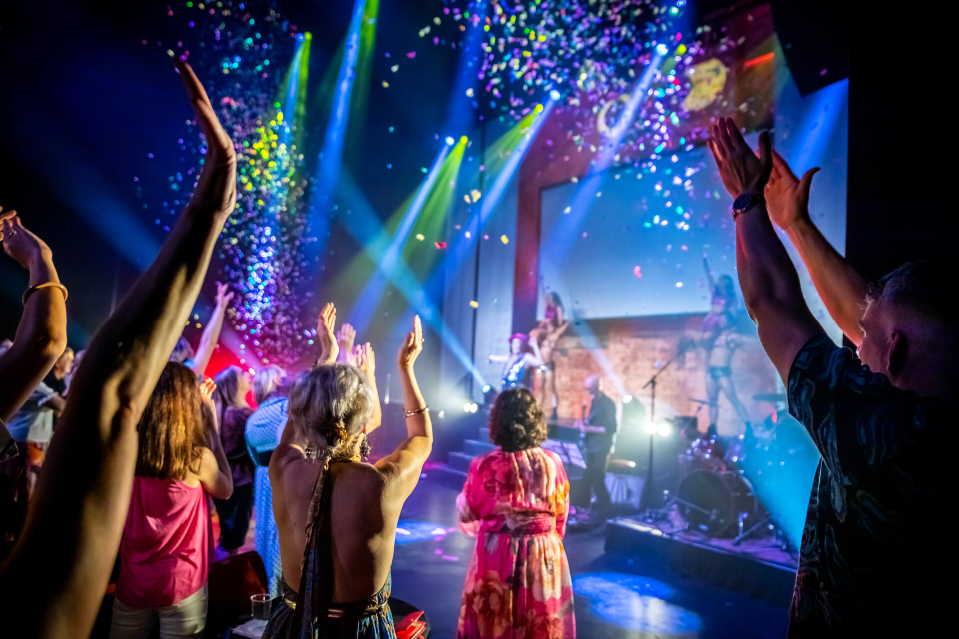 A crowd of people with raised hands enjoy a lively Vale Peter Roy concert as colorful confetti falls, bright stage lights shine, and musicians perform on stage in the background.